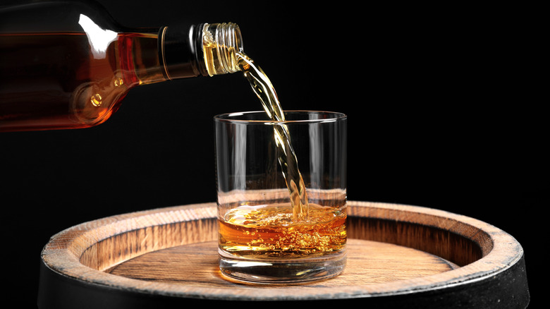 A bottle of bourbon pouring into a rocks glass atop a barrel