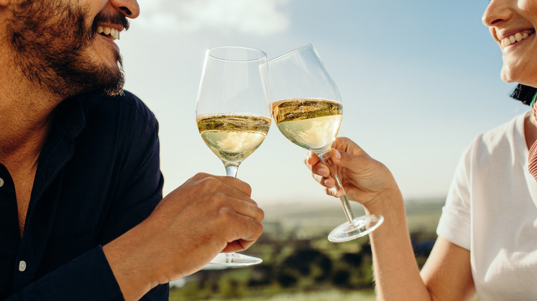Two people sitting outdoors toasting with glasses of white wine.