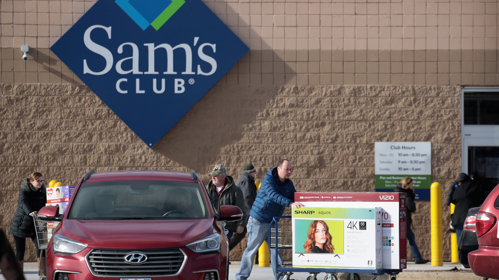 Man pushing cart with two television boxes outside of Sam's club. Maroon Hyundai parked nearby. 