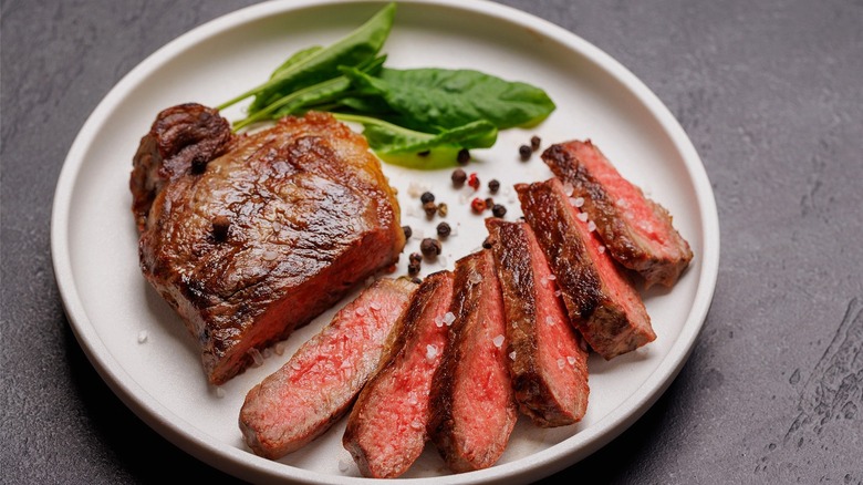 A sliced medium-rare steak served with greens and whole peppercorns on a white plate.
