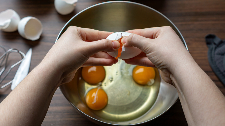 Hands cracking egg over metal bowl on wooden table with shells, napkin, and kitchen tools below