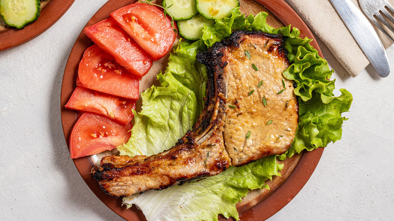 A browned pork chop on a ceramic plate with lettuce, sliced tomato, and cucumber