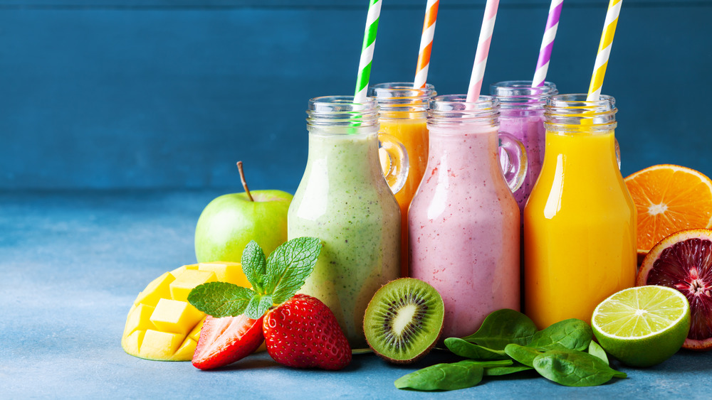 5 protein shakes of different colors with straws, fresh fruit, on gray surface 