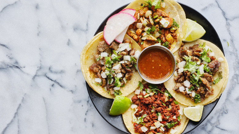 A plate with four Mexican style tacos, lime slices, radish and salsa 