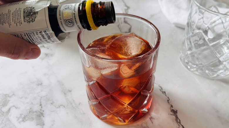 hand holding bitters bottle above a drinking glass filled with ice and brown liquid