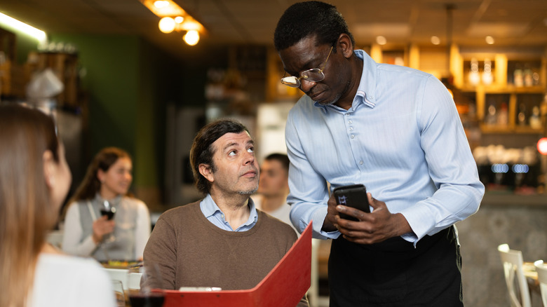 Man ordering wine at restaurant holds menu and looks at waiter with inquisitive expression