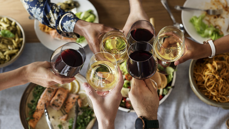 People toasting glasses of red and white wine over a table of plates of food like pasta, salads, and salmon