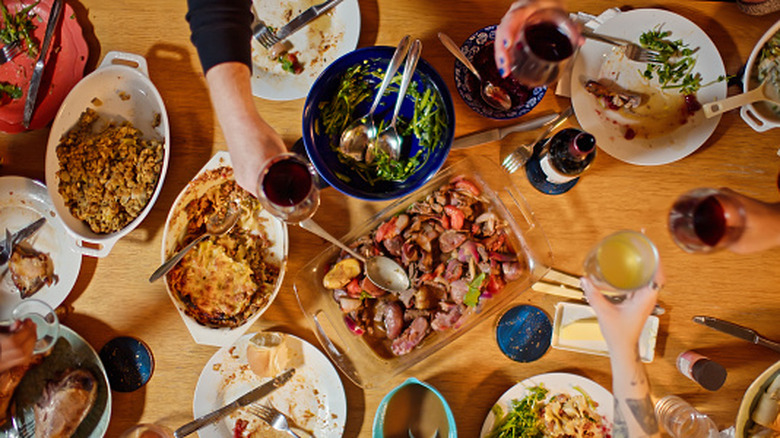 Aerial view of wooden table with people raising wine glasses over food, plates, and casserole dishes