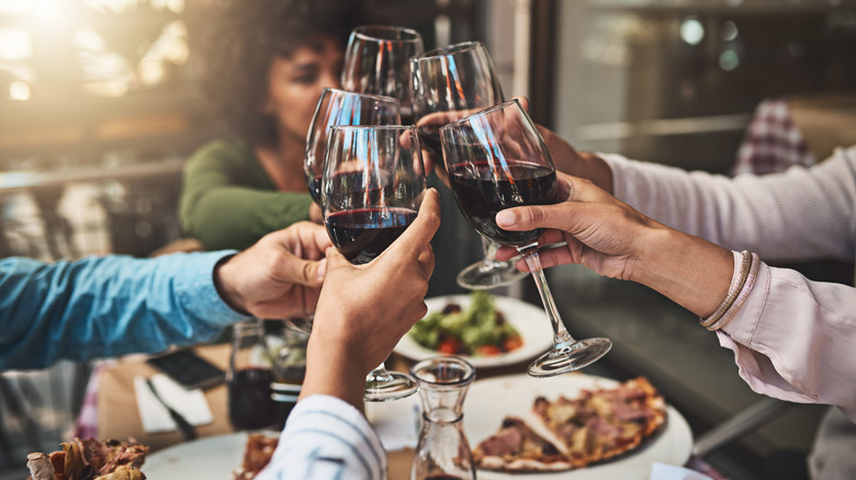 Hands toasting with glasses of red wine over table with plates of pizzas and salads