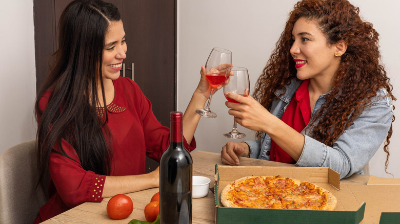 Two women clinking wine glasses together at table with a box of takeout pizza