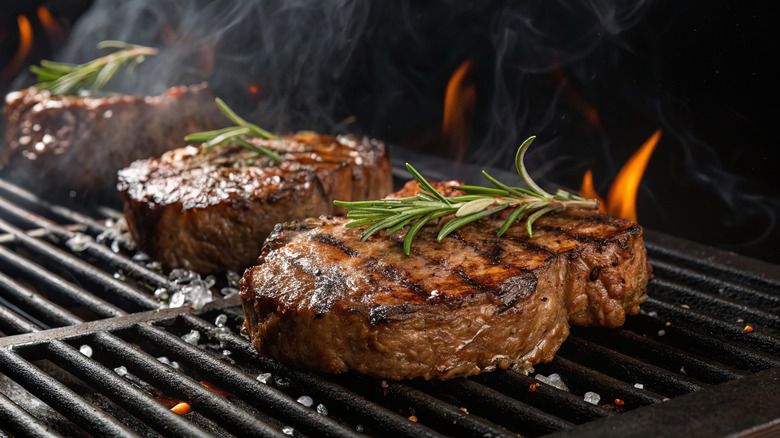 Three steaks cooking on a smoky grill with fresh herbs on top