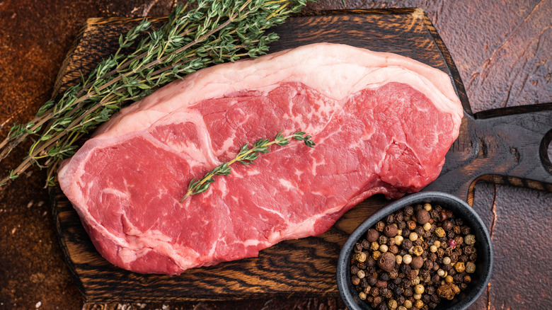 Well-marbled cut of raw NY strip steak on cutting board with herbs and seasonings