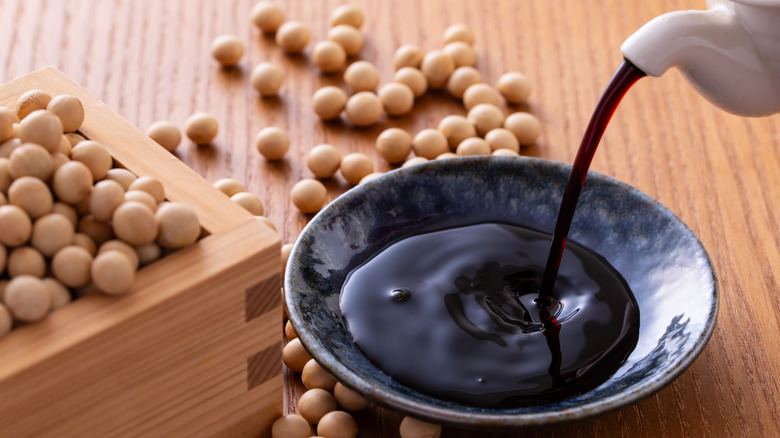 Pouring soy sauce into a bowl near a box of soybeans as more soybeans are scattered on the table.