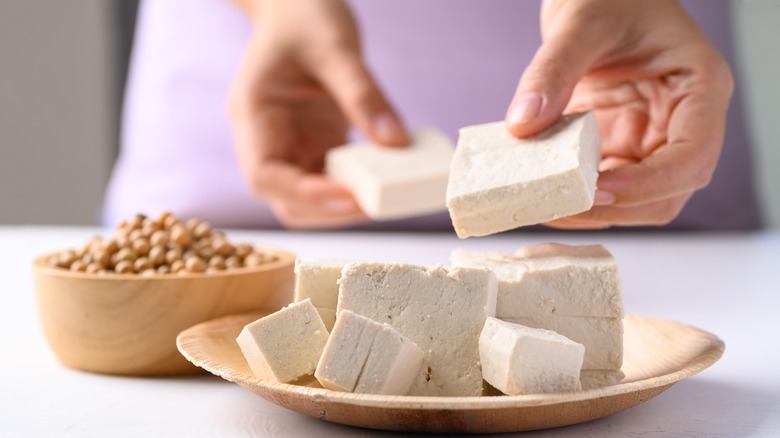 Person breaking apart a block of tofu on wooden plate next to wood bowl of soy beans