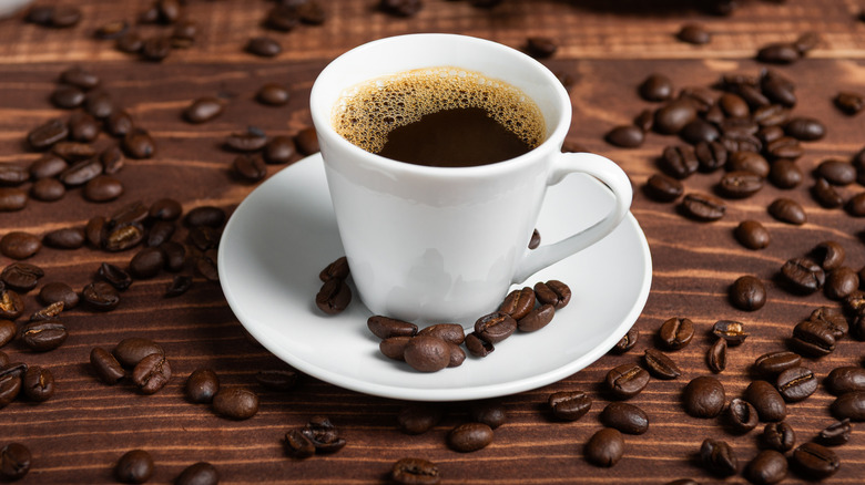 A mug of coffee sits on a small plate on a wooden surface, surrounded by scattered coffee beans.