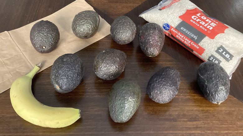 Eight avocados on a table with a banana, a paper bag, and a bag of rice