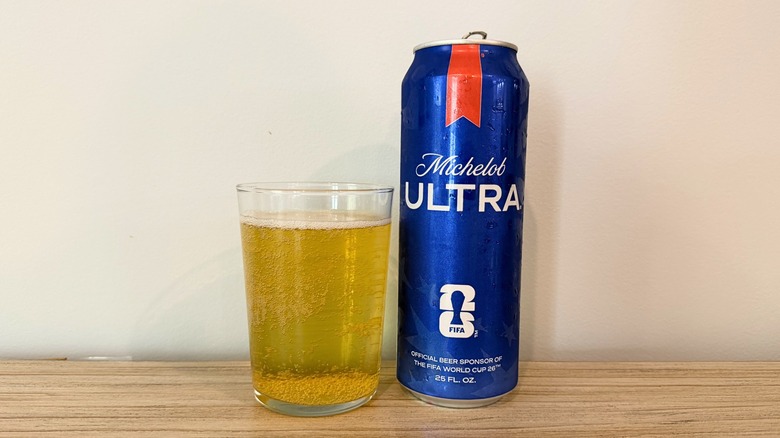 A glass of Michelob Ultra beside a tall blue Michelob Ultra can, showing a pale golden beer with bubbles