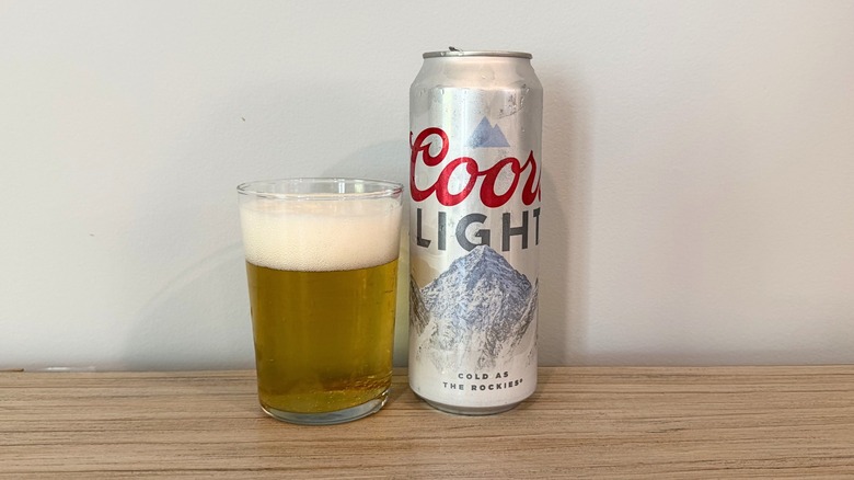 A glass of Coors Light beside a silver Coors Light can, filled with pale golden beer topped with a thick, creamy foam head