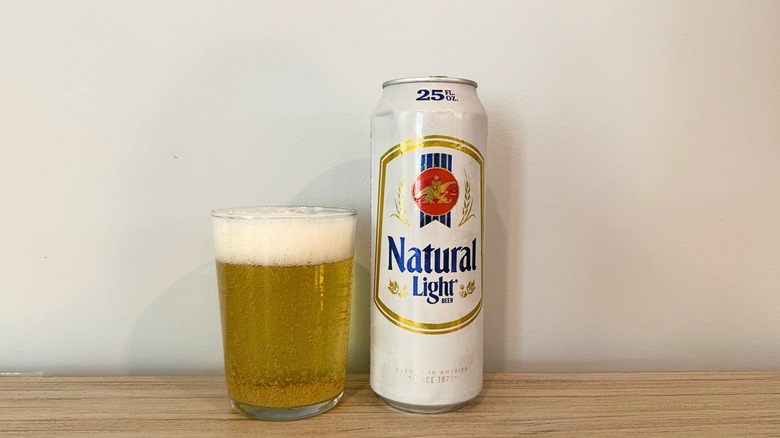 A glass of Natural Light next to a tall white Natural Light can, filled with bubbly pale beer and topped with a thick foam head