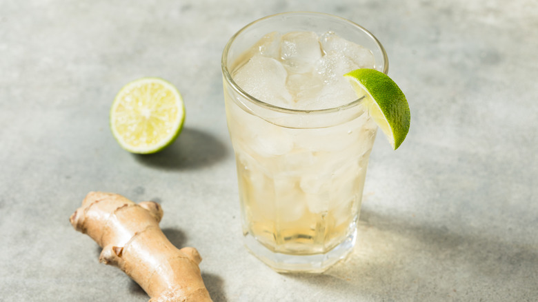 glass of ginger ale with a slice of lime on the edge of the glass and ginger root and half a lime resting nearby