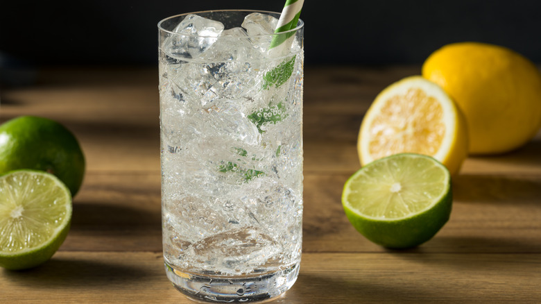 glass of lemon lime soda with a green and white striped straw and fresh limes and lemons in background