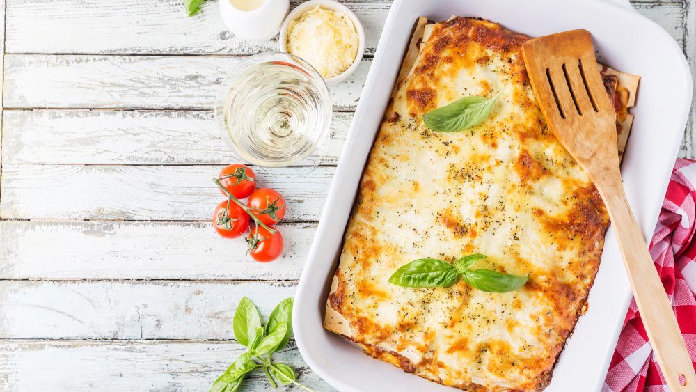 Lasagna in a baking dish next to basil, cheese, tomatoes and a glass of white wine