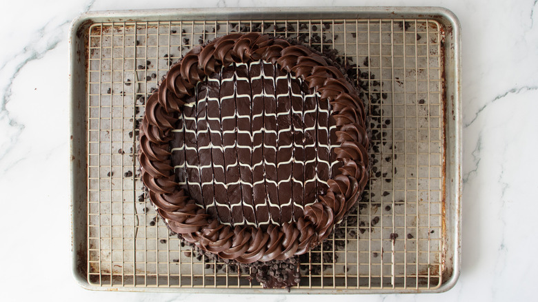chocolate-frosted cake with a wavy white drizzle, mini chips, and a chocolate frosting border on a metal rack