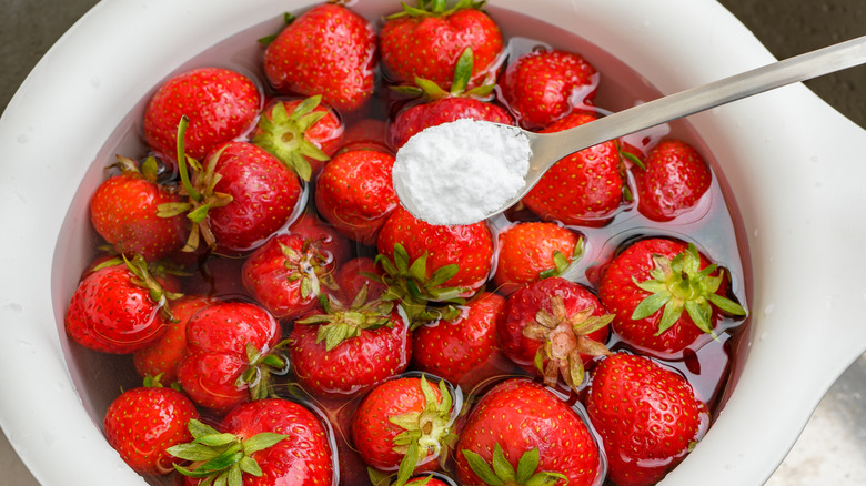 Spoon of baking soda above bowl of strawberries in water.