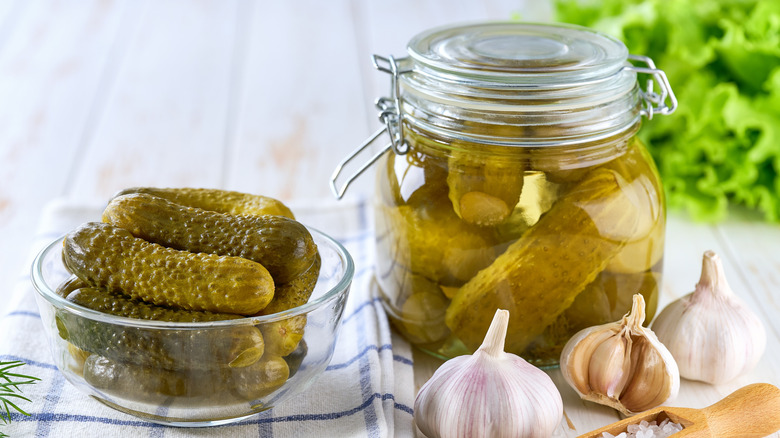 A bowl and jar of pickles on a kitchen table surrounded by herbs and spices