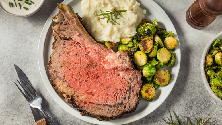 A cut of pinkish prime rib on a plate with mashed potatoes and Brussels sprouts