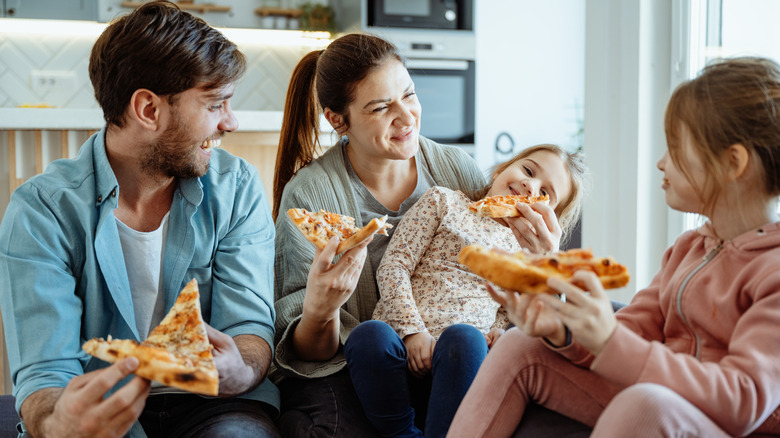 a family of four with two young daughters laughing and eating pizza in living room