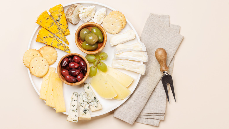 A white platter with assorted cheeses and crackers, green grapes, and red and green olives with a beige cloth napkin and wooden fork on a beige counter