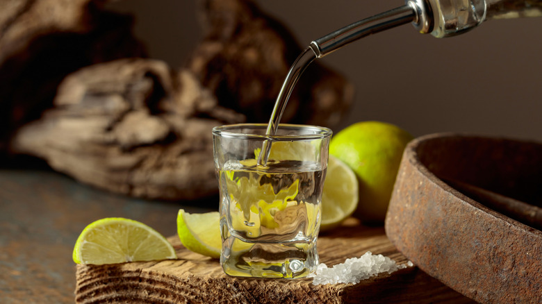 A pour of a shot glass of tequila on a wooden board with lime and salt
