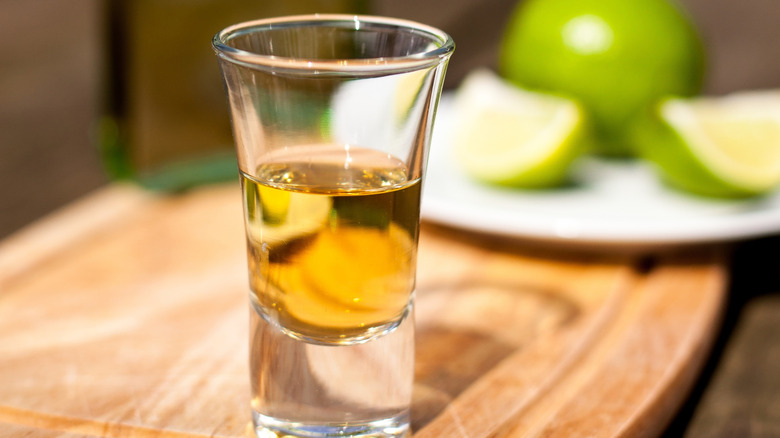 A shot glass filled with amber tequila on a wooden board, blurred limes in the background