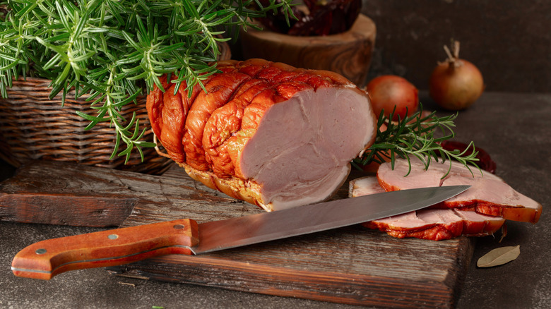 Smoked ham on wooden cutting board with knife and basket of rosemary
