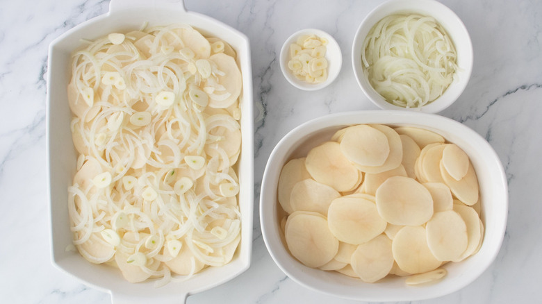Potatoes, onion, and garlic layered in casserole dish next to bowl of garlic, onions, and more potatoes