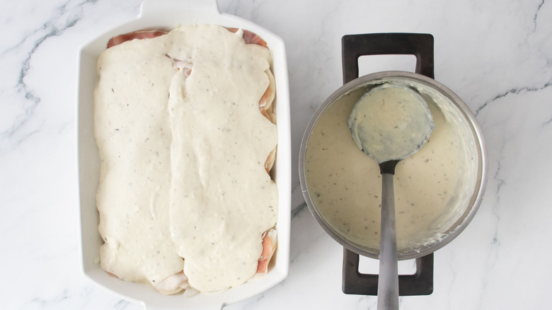 Layer of creamy sauce in casserole dish next to sauce and ladle in pan
