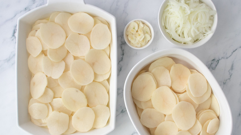 Potato slices layered in casserole dish next to bowl of garlic, onions, and more potato slices