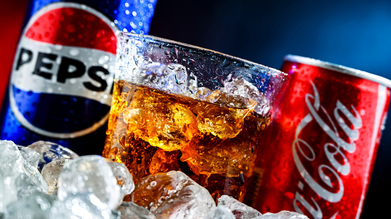 Cola in glass, can of Pepsi blurred on left, can of Coke blurred on right