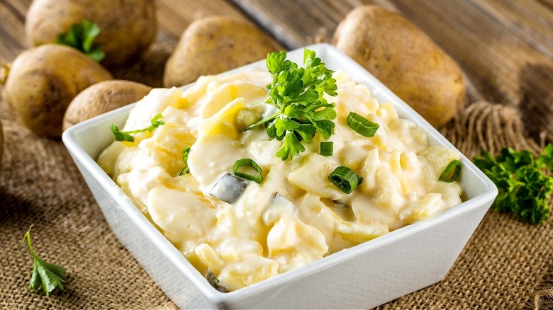 potato salad in a white dish surrounded by raw potatoes and parsley