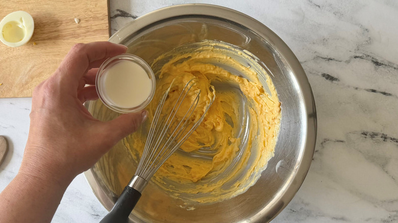 Hand holding small bowl of cream over egg salad in bowl with whisk