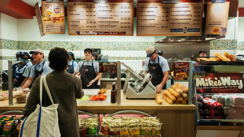 Employees stand behind service counter in a Jersey Mike's with a menu and a person facing the counter