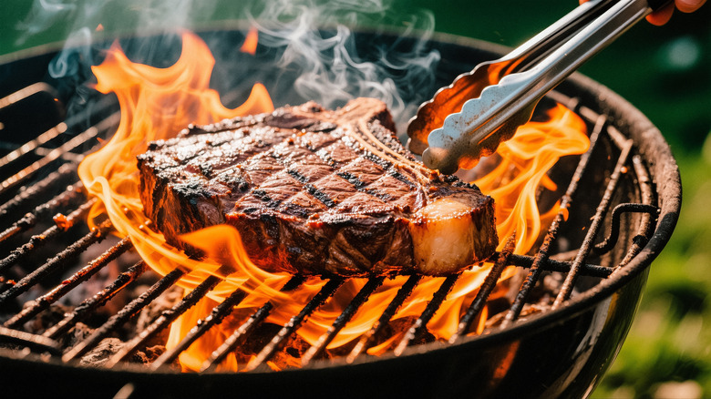 Tongs adjusting steak on a flaming grill.