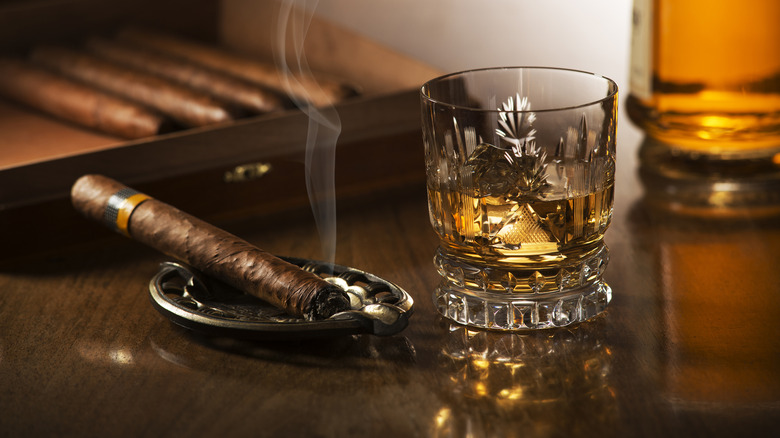 A half-full tumbler of whiskey next to smoking cigar on ashtray on table.