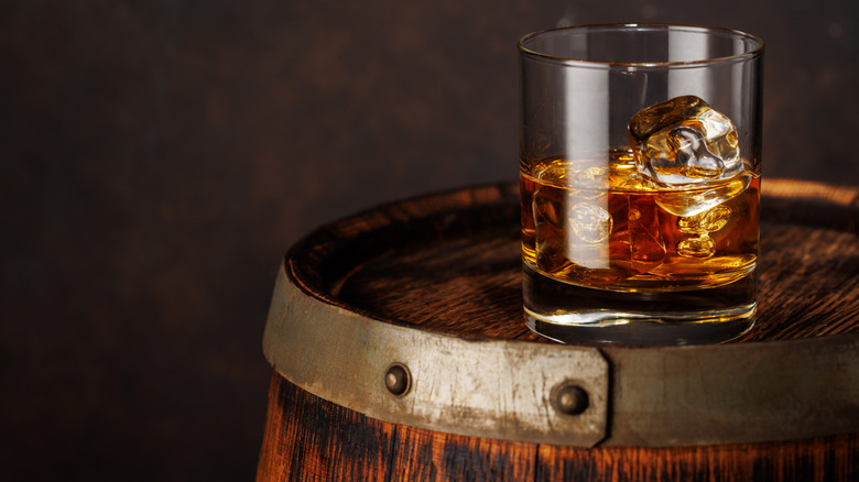 Glass of whiskey with ice sitting on top of aging barrel.