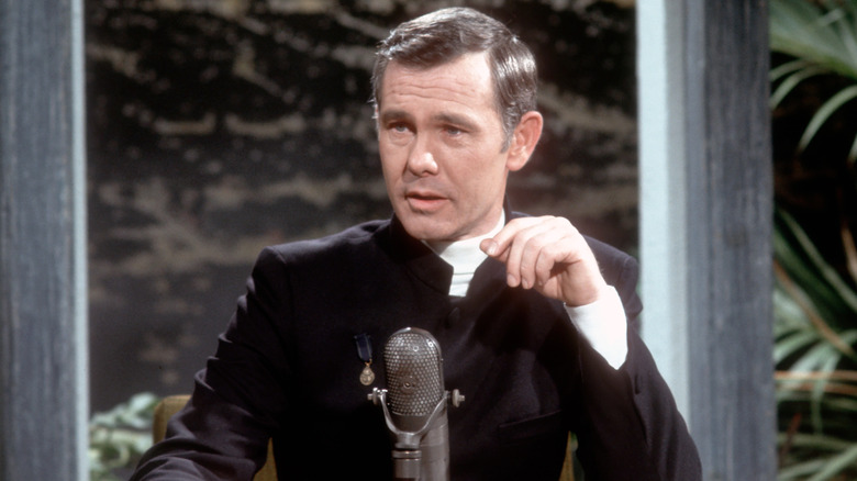 Johnny Carson at his host desk in a black suit and white quarter-neck shirt