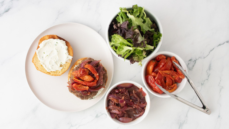 Lamb burger topped with roasted tomatoes next to bowls of toppings