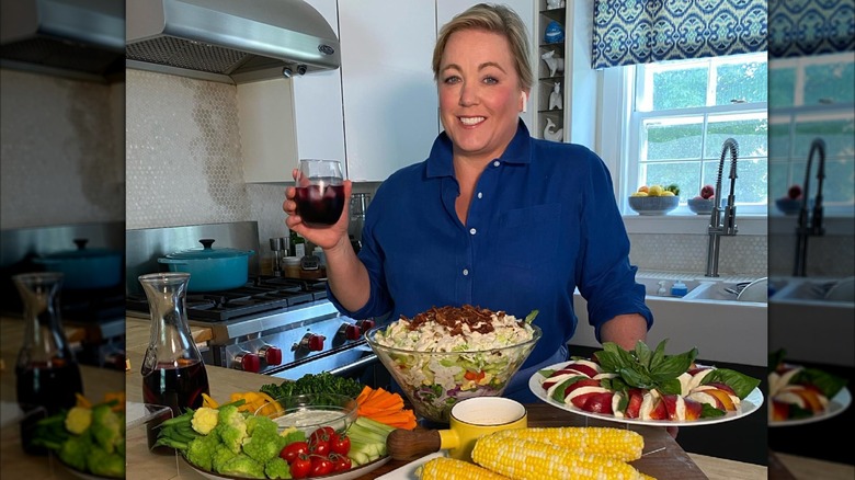 Julia Collin Davison smiles while holding a glass of wine in front of a healthy food spread