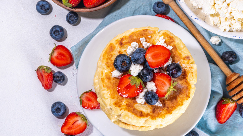 Pancakes topped with blueberries, strawberries, and cottage cheese next to a honey stick and more fruit.