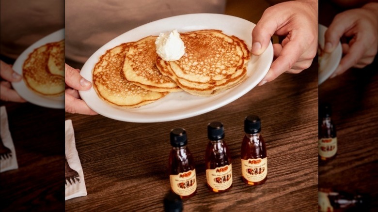Hands holding a plate of Cracker Barrel pancakes topped with butter, next to three mini bottles of syrup